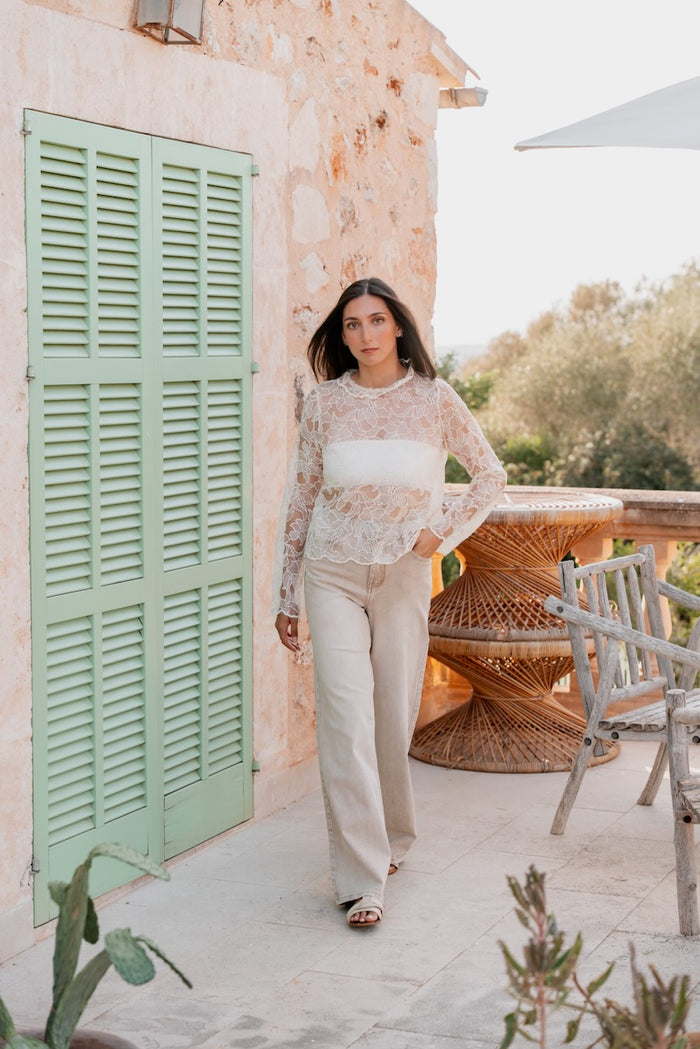 La tenue se compose d'une blouse à manches longues en dentelle blanche transparente à motifs floraux, dotée d'un col rond et superposée à un bandeau blanc. Elle est associée à un pantalon long à coupe large en toile beige clair, tombant sur des sandales plates pour une silhouette fluide.
