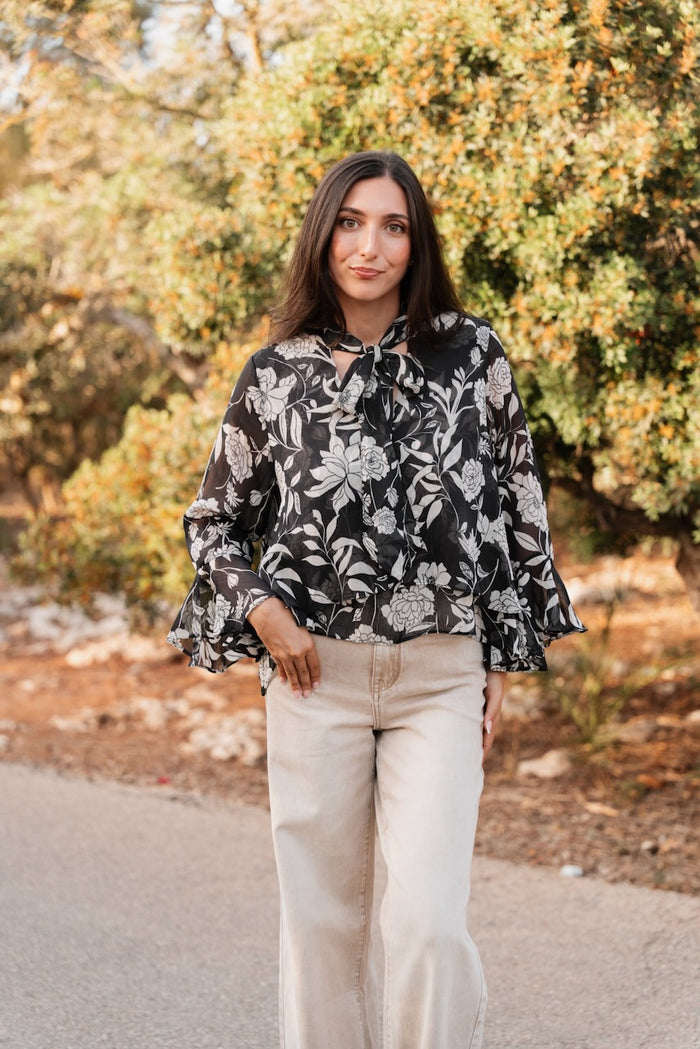 Blouse fluide à manches longues évasées terminées par des volants, de couleur noire ornée de motifs floraux et de feuillages blancs. L'encolure est caractérisée par un col lavallière noué en un nœud souple. La coupe est ample et le tissu semi-transparent. Elle est associée à un jean large de couleur écru.