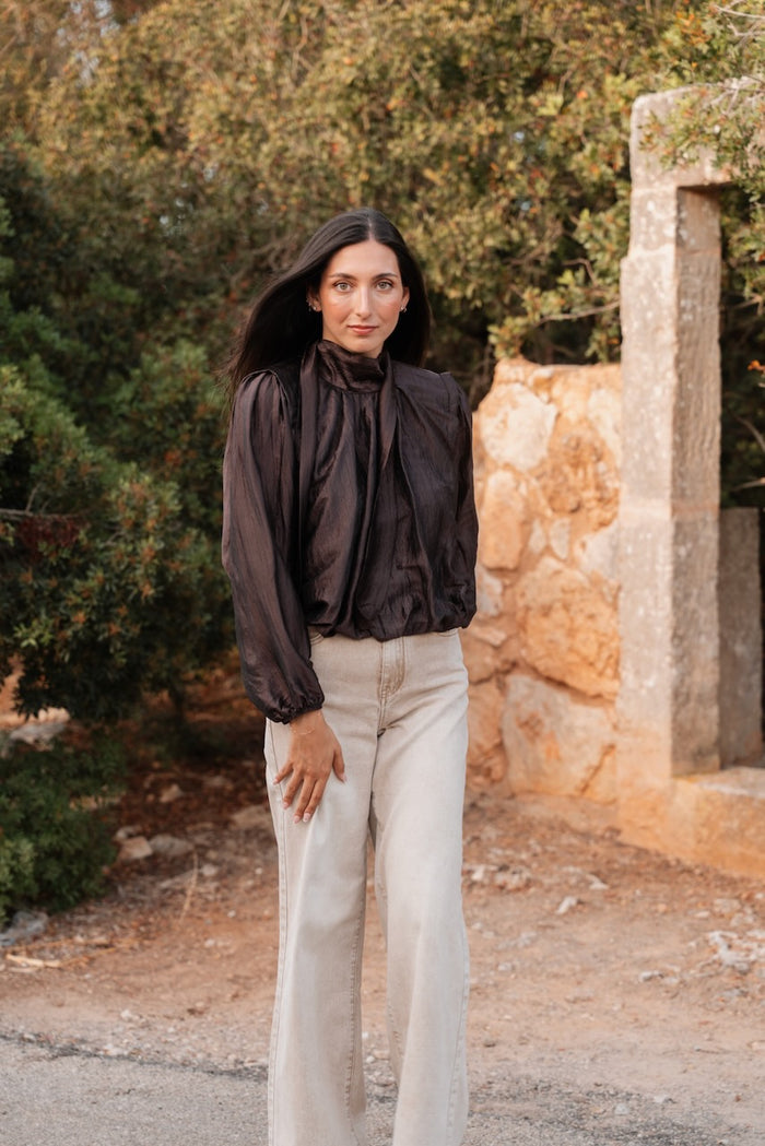 Une blouse à col montant en tissu texturé et brillant marron foncé est portée sur un jean large de couleur beige délavé. Le haut blousant présente une écharpe intégrée drapée sur le devant, des manches longues bouffantes aux poignets élastiques et une taille élastiquée créant un effet blousonné. L'association se fait avec un jean à jambe large, de coupe droite et à passants de ceinture.