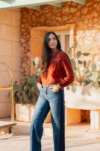 Blouse fluide boutonnée de couleur brique, confectionnée dans un tissu à texture visible. Elle se distingue par une grande collerette volantée ornée de broderies blanches contrastées autour de l'encolure fendue. Les manches longues sont bouffantes, froncées aux emmanchures et aux poignets. La blouse est boutonnée sur le devant avec des boutons assortis et est associée à un jean bleu délavé et une ceinture en cuir marron.