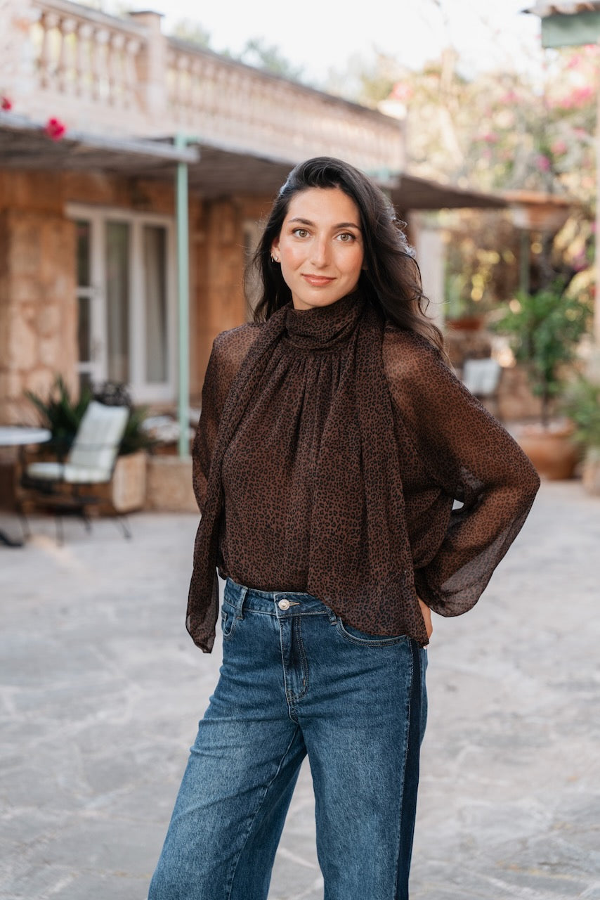 Une blouse fluide et semi-transparente de couleur marron foncé arbore un imprimé léopard dense. Le vêtement est doté d'un col montant qui se prolonge en une cravate lavallière lâche à l'avant. Ses manches sont longues et bouffantes, se terminant par des poignets élastiques froncés. Elle tombe amplement sur le corps.