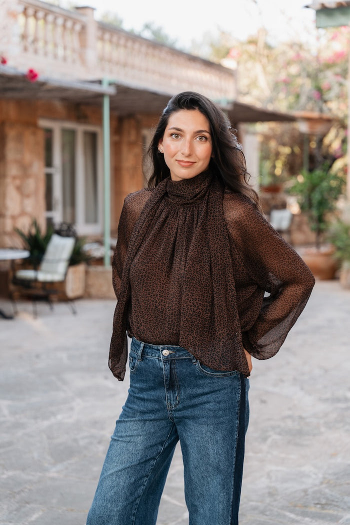 Une blouse fluide et semi-transparente de couleur marron foncé arbore un imprimé léopard dense. Le vêtement est doté d'un col montant qui se prolonge en une cravate lavallière lâche à l'avant. Ses manches sont longues et bouffantes, se terminant par des poignets élastiques froncés. Elle tombe amplement sur le corps.