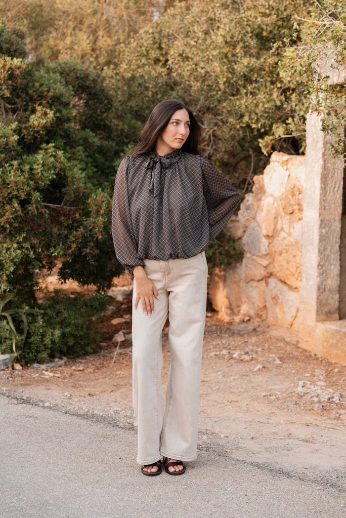 Une femme debout en extérieur porte un ensemble composé d'une blouse et d'un pantalon. La blouse fluide est de couleur noire à motifs de petits carreaux fins et clairs, avec un col montant froncé orné d'un nœud lavallière. Elle présente une coupe blousante à la taille et des manches longues et amples resserrées aux poignets. Le pantalon est en denim de coton beige clair, de coupe large et droite jusqu'au sol, avec une taille haute apparente.