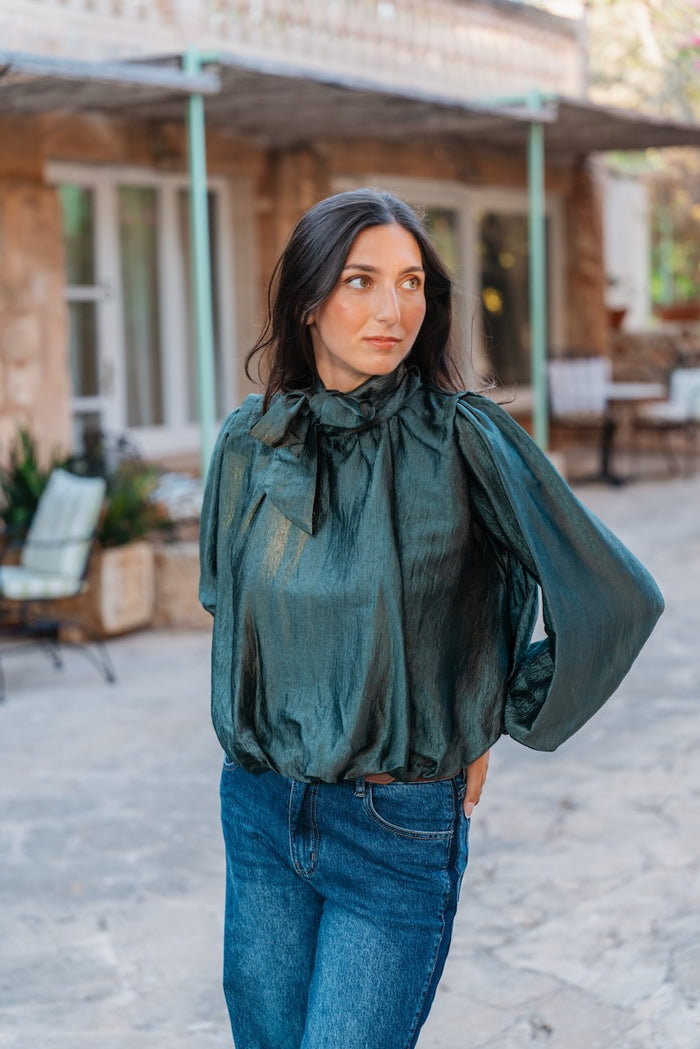 Une blouse blousante et fluide, confectionnée dans un tissu lamé vert émeraude texturé et brillant, présente un col montant pussy-bow noué en un grand nœud papillon à l'avant. Elle est dotée de manches longues et très volumineuses, froncées, et d'un ourlet élastiqué à la taille qui crée un effet blouson. Une fine ceinture en cuir marron est visible au-dessus d'un jean en denim bleu moyen à coupe droite et légèrement usé.