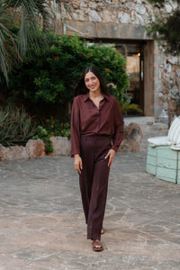 L'image présente une femme debout portant un ensemble coordonné. Le haut est une chemise bordeaux à manches longues avec de fines rayures verticales argentées, ornée de discrets points brillants incrustés. La chemise, au col classique, est rentrée dans un pantalon large marron foncé uni à taille haute. Le pantalon a une coupe fluide et tombe droit.Elle complète la tenue avec des sandales plates marron clair.