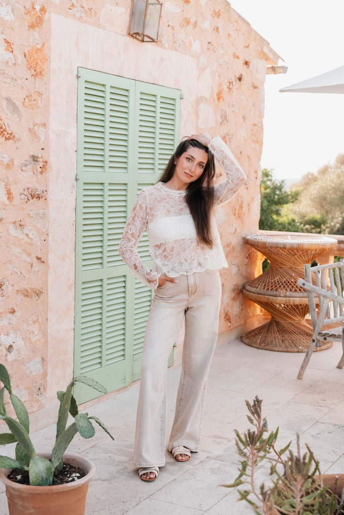 Une tenue de deux pièces composée d'un haut en dentelle blanche et d'un pantalon large beige clair est présentée sur une femme debout. Le haut est en dentelle florale translucide à manches longues, avec une encolure ronde simple. Il est porté sur un haut de bandeau blanc opaque, créant un effet de superposition visible à travers la dentelle. Le pantalon est en denim de coton beige clair, avec une coupe à jambes larges et droites, une taille haute et des poches. La silhouette d'ensemble est fluide, le haut n