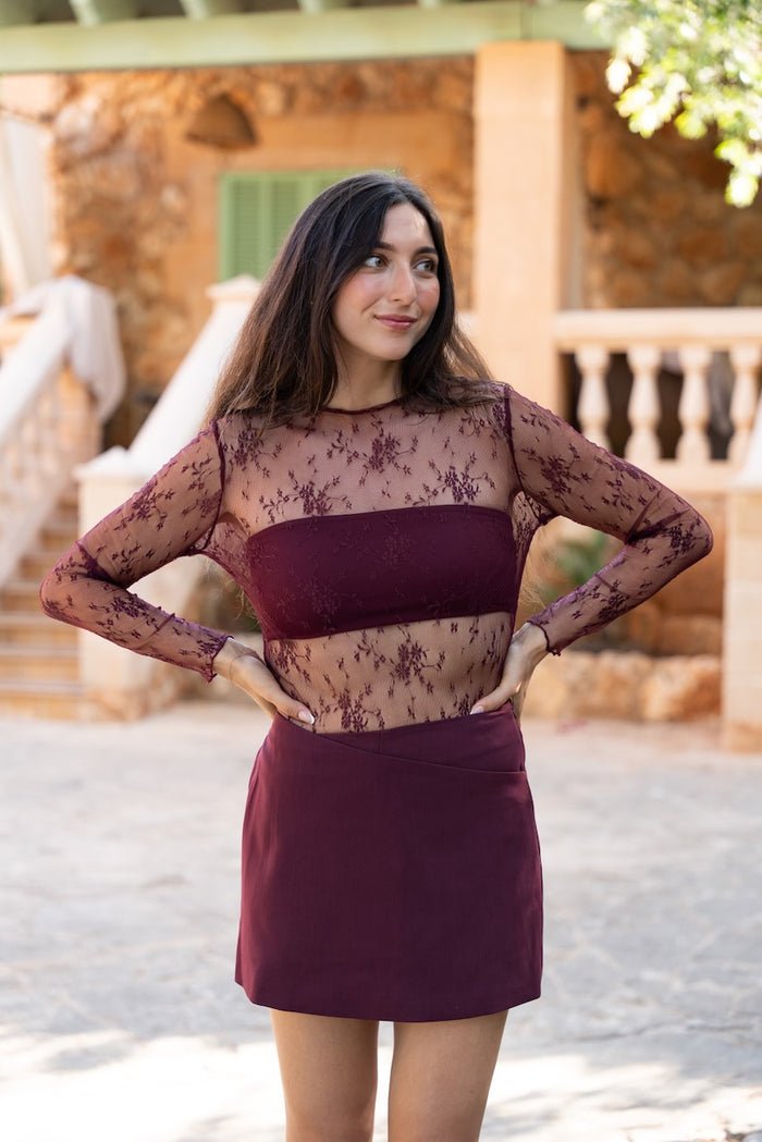 Cet ensemble coordonné de couleur bordeaux est composé d'un haut et d'une jupe. Le haut est une blouse à manches longues en maille de dentelle florale fine et transparente, dotée d'une encolure ronde simple. Sous la dentelle, un bandeau opaque assorti est intégré pour couvrir le buste, tandis que la transparence est visible sur les bras, le décolleté supérieur et la taille. La jupe coordonnée est une jupe crayon courte et opaque, tombant juste au-dessus du genou.
