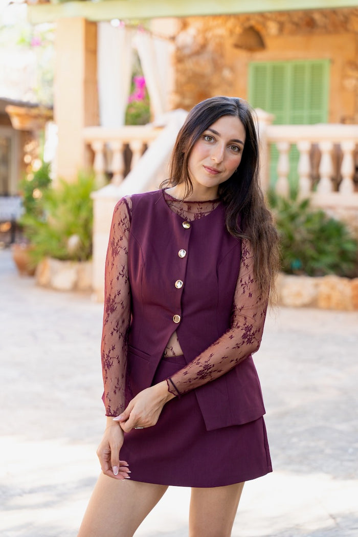 Un ensemble assorti bordeaux composé d'un gilet ajusté et d'une mini-jupe droite. Le gilet sans manches en tissu uni comporte quatre gros boutons dorés décoratifs et un ourlet avant asymétrique. Il est superposé à un haut en dentelle fine et translucide à manches longues, visible à l'encolure ronde et sur les bras avec son motif floral. La jupe assortie est courte,s'arrêtant mi-cuisse.