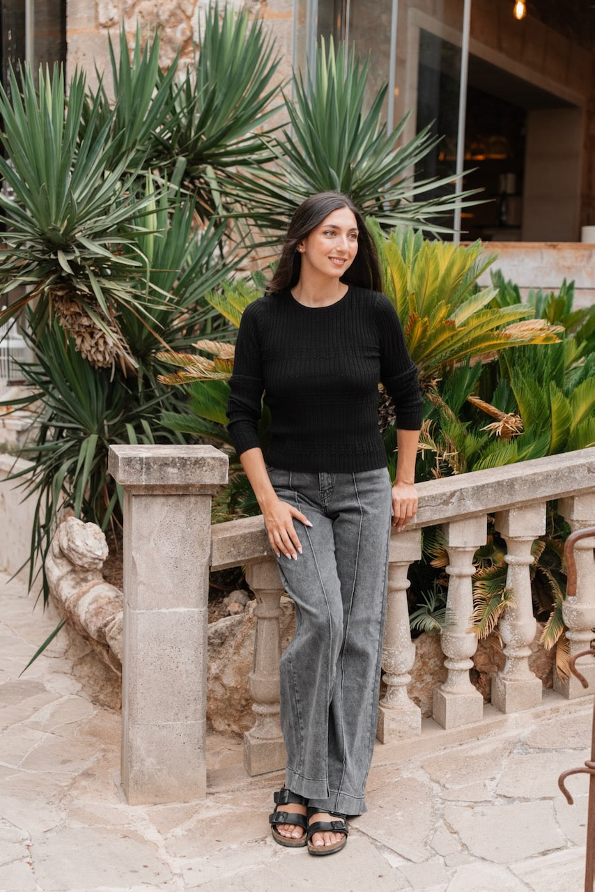 Un pull ajusté noir à manches longues et encolure ronde, fabriqué en maille côtelée avec une fine texture verticale,possède des poignets et un ourlet légèrement plus larges et texturés. Il est associé à un jean délavé gris moyen à coupe large, caractérisé par des coutures centrales marquées et des surpiqûres visibles sur le devant des jambes.