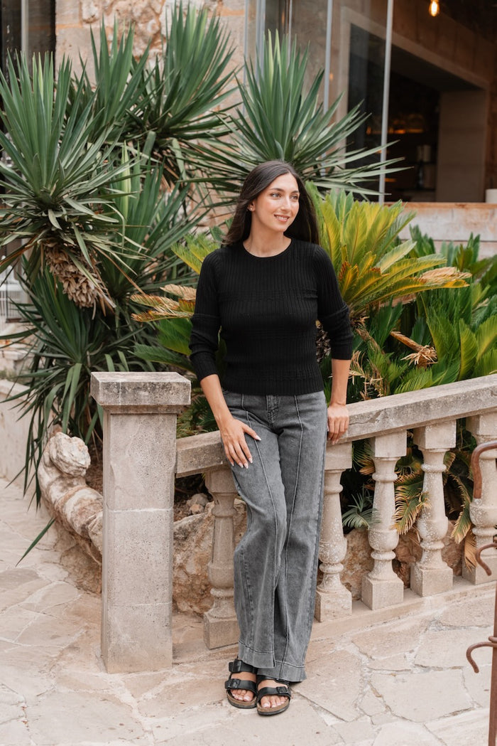 Un pull ajusté noir à manches longues et encolure ronde, fabriqué en maille côtelée avec une fine texture verticale,possède des poignets et un ourlet légèrement plus larges et texturés. Il est associé à un jean délavé gris moyen à coupe large, caractérisé par des coutures centrales marquées et des surpiqûres visibles sur le devant des jambes.