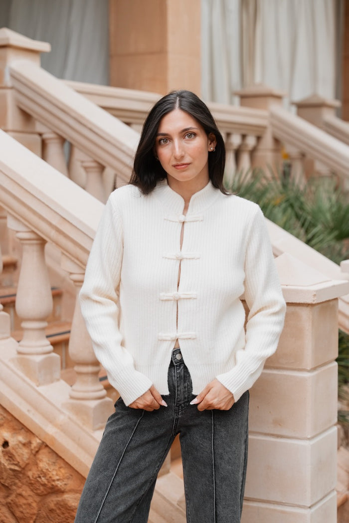 Un cardigan court en tricot côtelé de couleur crème, doté d'un col mao montant et de manches longues avec poignets côtelés. Le vêtement est structuré et se ferme au centre sur le devant par quatre brandebourgs en tissu assorti, laissant un petit espace visible entre les attaches. La longueur du cardigan est courte, arrivant au niveau de la hanche haute.