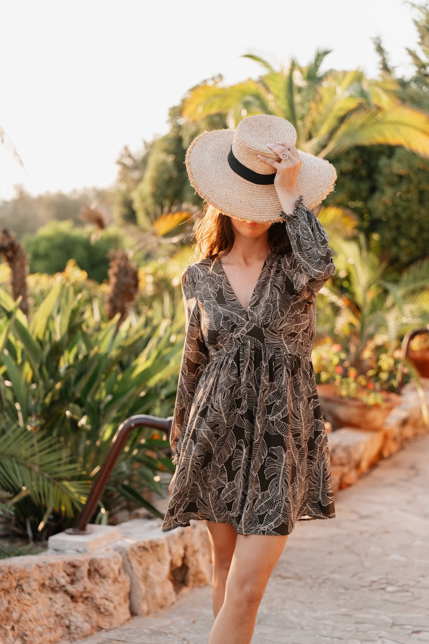 Robe courte fluide de couleur noire avec un imprimé intégral de feuilles de palmier stylisées en gris clair. La silhouette est de style empire avec un décolleté en V plongeant et une taille haute froncée juste sous la poitrine qui se prolonge en une jupe légèrement évasée arrivant au-dessus du genou. Les manches sont longues, fluides et présentent des poignets élastiqués. Le tissu est léger et texturé de manière unie par le motif.
