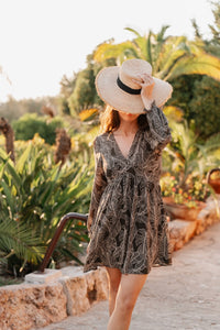 Robe courte fluide de couleur noire avec un imprimé intégral de feuilles de palmier stylisées en gris clair. La silhouette est de style empire avec un décolleté en V plongeant et une taille haute froncée juste sous la poitrine qui se prolonge en une jupe légèrement évasée arrivant au-dessus du genou. Les manches sont longues, fluides et présentent des poignets élastiqués. Le tissu est léger et texturé de manière unie par le motif.