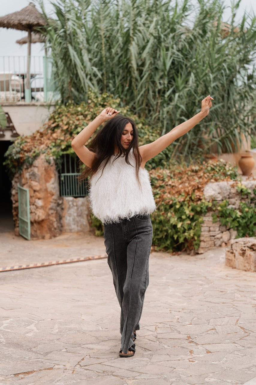 Une femme en marche porte un haut sans manches couleur crème au col rond, entièrement couvert de longues franges pelucheuses qui créent une texture dense et duveteuse. Le haut a une coupe ample et s'arrête juste au-dessus des hanches.Elle est vêtue d'un jean large de couleur gris foncé délavé qui tombe sur ses pieds, caractérisé par des coutures verticales décoratives le long des jambes. Aux pieds, elle porte des sandales plates noires à double bride.