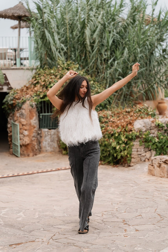 Une femme en marche porte un haut sans manches couleur crème au col rond, entièrement couvert de longues franges pelucheuses qui créent une texture dense et duveteuse. Le haut a une coupe ample et s'arrête juste au-dessus des hanches.Elle est vêtue d'un jean large de couleur gris foncé délavé qui tombe sur ses pieds, caractérisé par des coutures verticales décoratives le long des jambes. Aux pieds, elle porte des sandales plates noires à double bride.