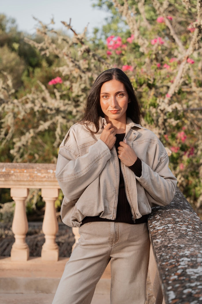 Un ensemble coordonné de couleur beige clair, composé d'une veste écourtée oversize et d'un pantalon à jambe large. La veste est confectionnée dans une toile de denim texturée et présente une silhouette volumineuse avec des manches larges resserrées aux poignets, un col chemise et une fermeture éclair argentée centrale. Elle est dotée de poches plaquées discrètes et d'un ourlet inférieur large et souple. Sous la veste, un pull en tricot fin marron foncé à col rond et manches longues est visible. Le pantalon