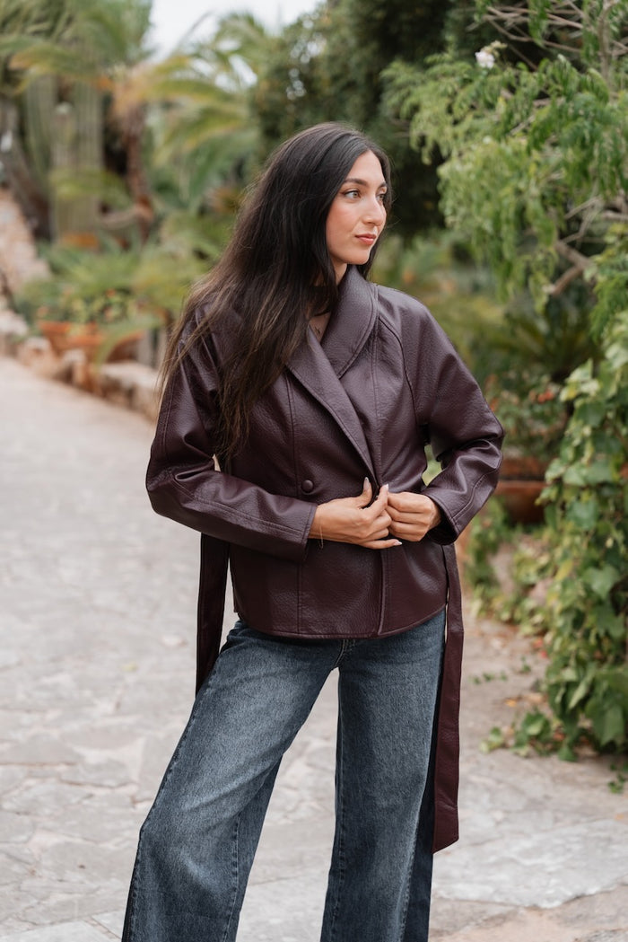 Veste d'extérieur de coupe droite en similicuir grainé de couleur bordeaux foncé, arrivant à mi-hanches. Elle présente un large col à revers et une fermeture croisée à double boutonnage sécurisée par deux boutons assortis. Une ceinture amovible du même matériau est nouée à la taille pour cintrer la silhouette. Les manches sont longues et droites, et aucun détail de poche n'est visible sur le devant.