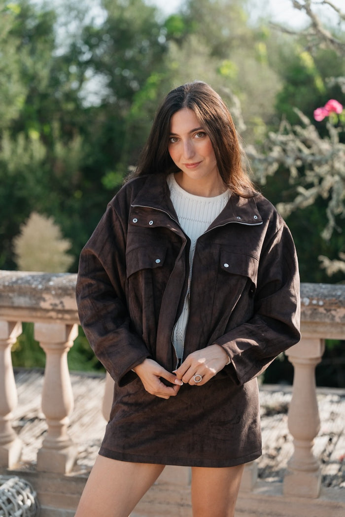 Un ensemble coordonné en suédine marron chocolat se compose d'une veste d'aviateur ample et d'une mini-jupe assortie.La veste volumineuse arbore un grand col convertible à boutons-pression argentés et une fermeture éclair centrale. Elle dispose d'épaules tombantes, de manches larges, d'un ourlet élastiqué et de poches poitrine plaquées avec des pressions décoratives. La mini-jupe droite et courte arrive à mi-cuisse, complétant la silhouette blousante de la veste. Un haut en tricot crème texturé est partiell